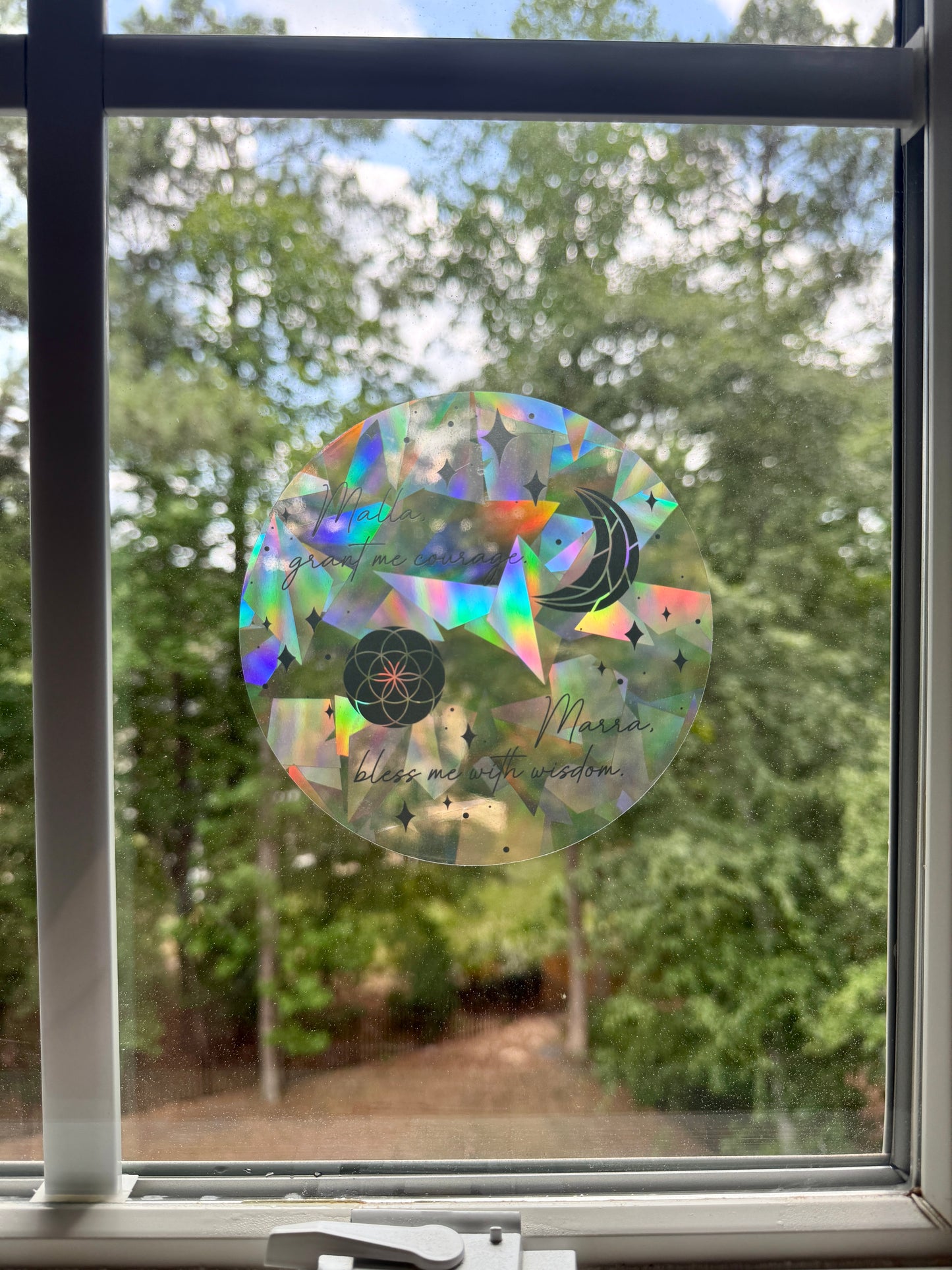 Decorative circular suncatcher inspired by The Road of Bones by Demi Winters with text 'Malla grant me courage. Marra bless me with wisdom' and moon and stars symbols on a window with a blurred outdoor background.