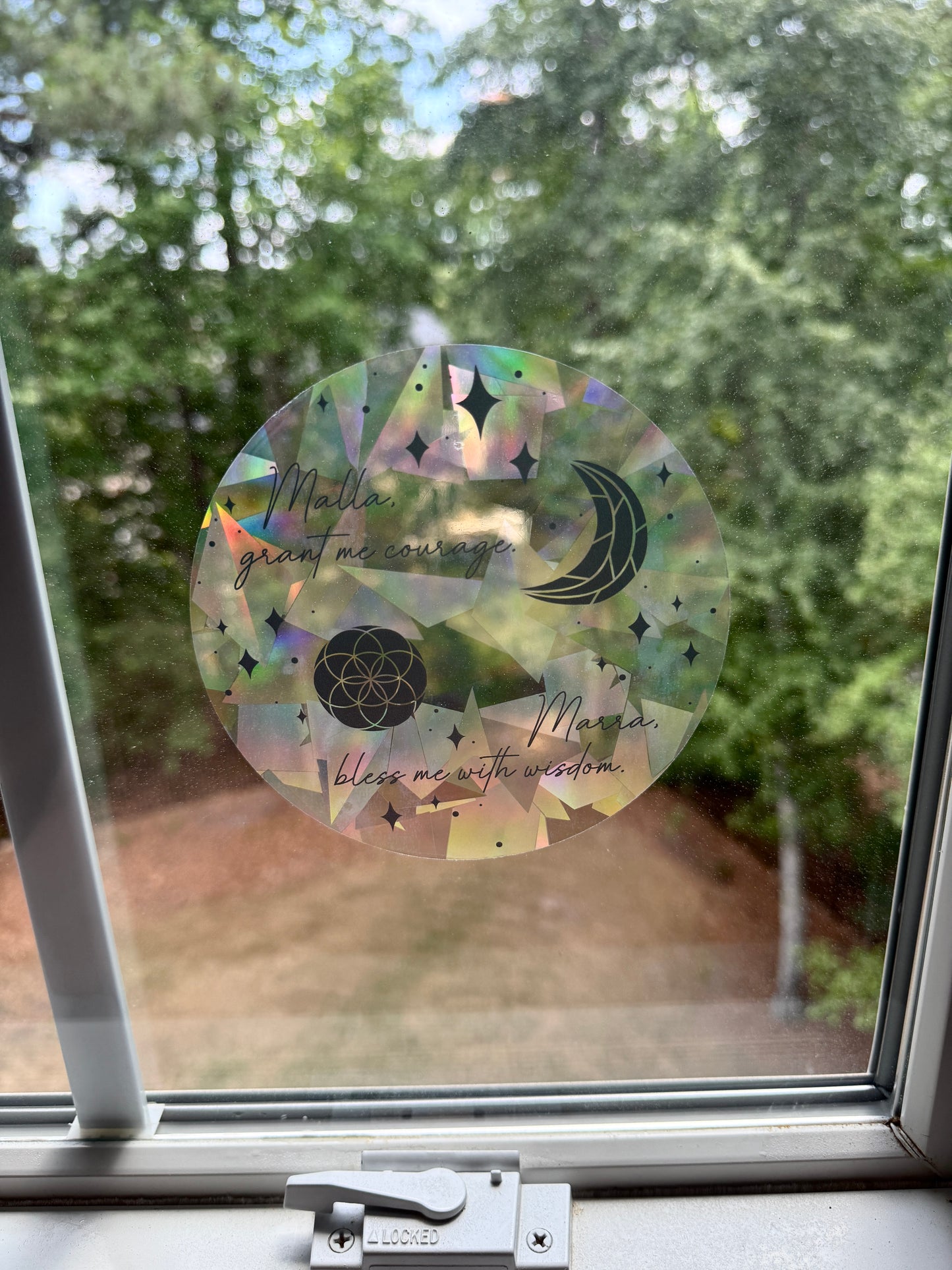 Decorative circular suncatcher inspired by The Road of Bones by Demi Winters with text 'Malla grant me courage. Marra bless me with wisdom' and moon and stars symbols on a window with a blurred outdoor background.