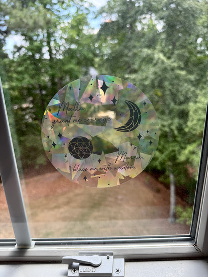 Decorative circular suncatcher inspired by The Road of Bones by Demi Winters with text 'Malla grant me courage. Marra bless me with wisdom' and moon and stars symbols on a window with a blurred outdoor background.