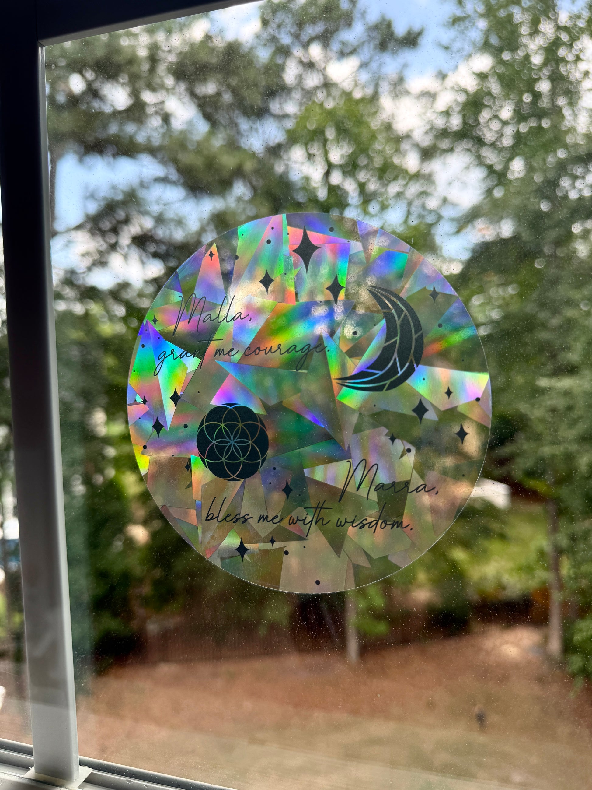 Decorative circular suncatcher inspired by The Road of Bones by Demi Winters with text 'Malla grant me courage. Marra bless me with wisdom' and moon and stars symbols on a window with a blurred outdoor background.