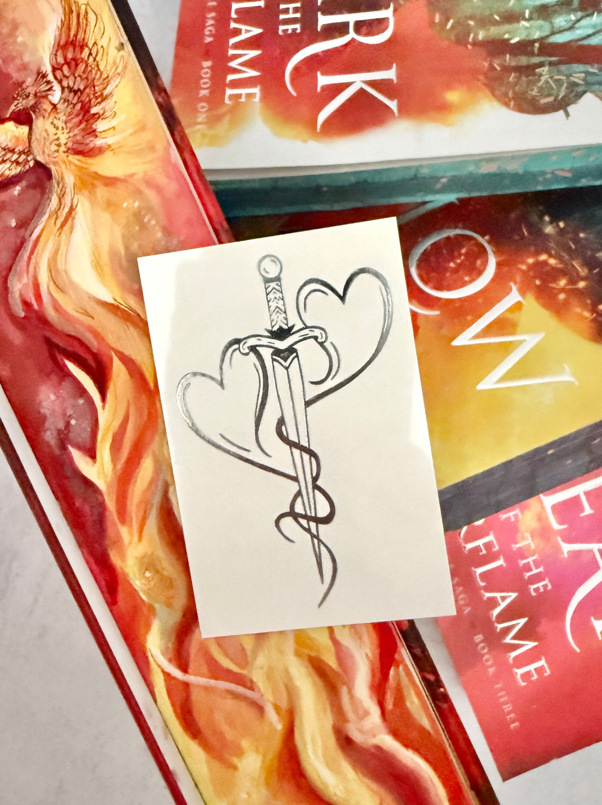 Two hearts forged into a dagger - the mating mark temporary tattoo resting on the Kindred's Curse Saga books by Penn Cole, one of which has painted edges with a phoenix and flames