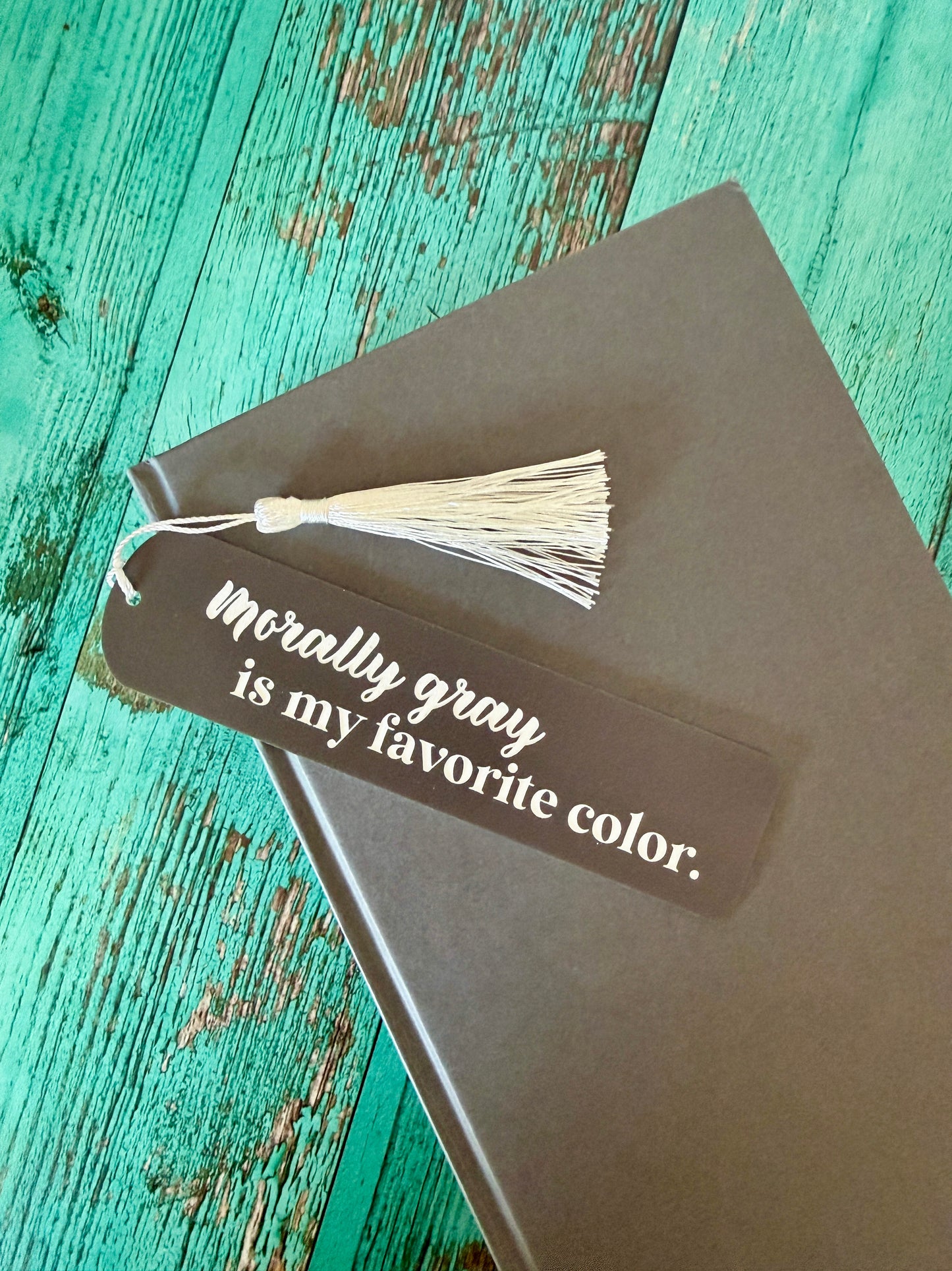 Book with a bookmark that reads "morally gray is my favorite color" on a teal wooden surface