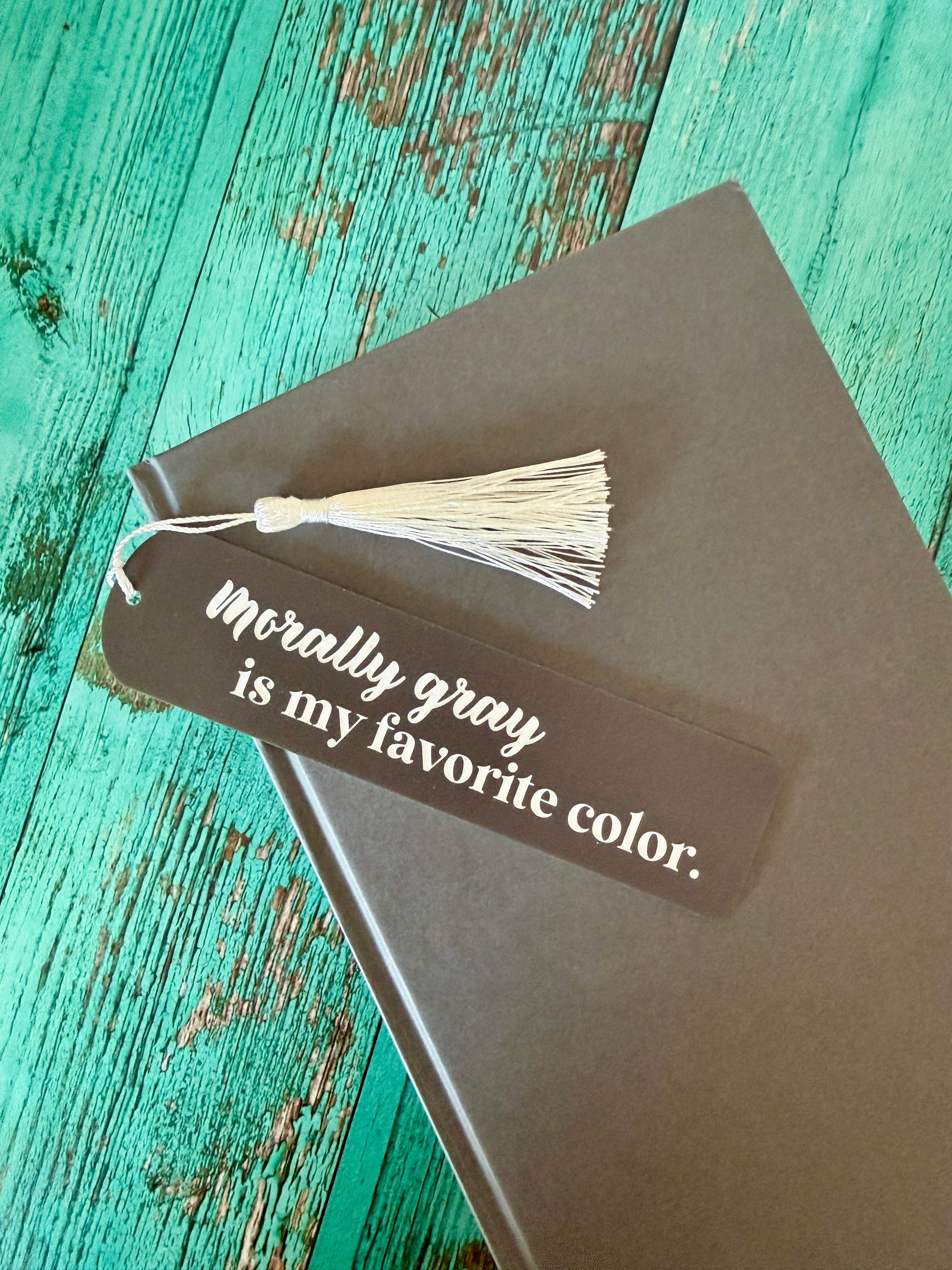 Book with a bookmark that reads "morally gray is my favorite color" on a teal wooden surface