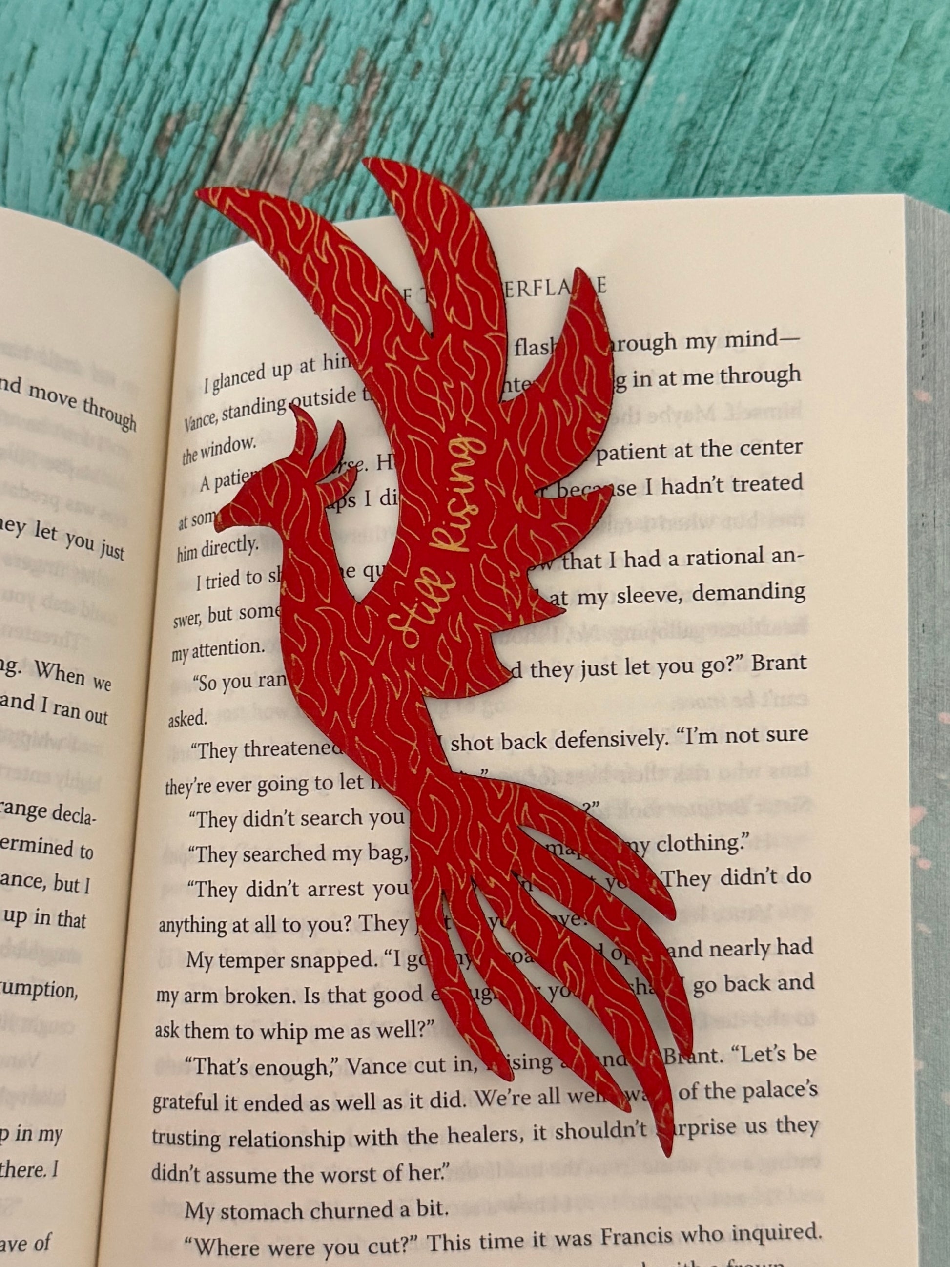 phoenix bookmark in red with gold engraving "still rising" sitting on open book