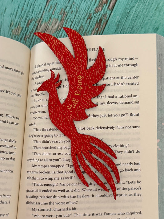 phoenix bookmark in red with gold engraving "still rising" sitting on open book