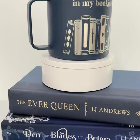 Navy stainless steel mug with 'in my bookish era' and books engraved around it, set on top of a stack of blue books.