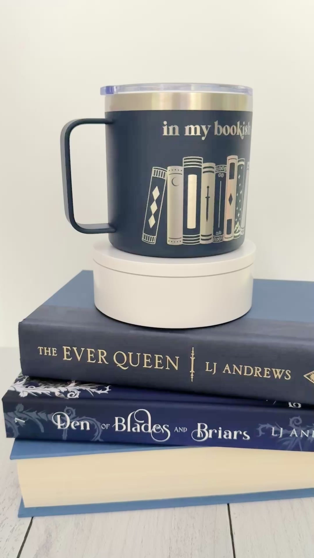 Navy stainless steel mug with 'in my bookish era' and books engraved around it, set on top of a stack of blue books.