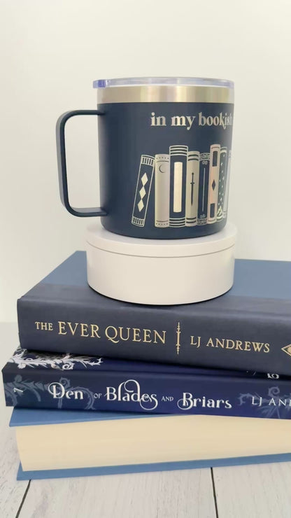 Navy stainless steel mug with 'in my bookish era' and books engraved around it, set on top of a stack of blue books.