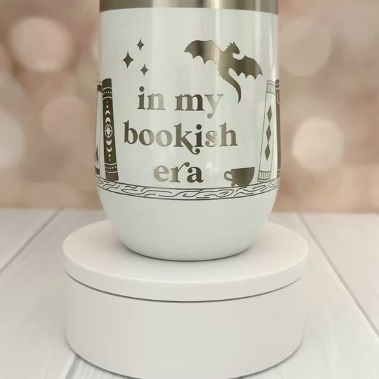 white wine tumbler engraved with "in my bookish era" and books on a bookshelf with a dragon flying above