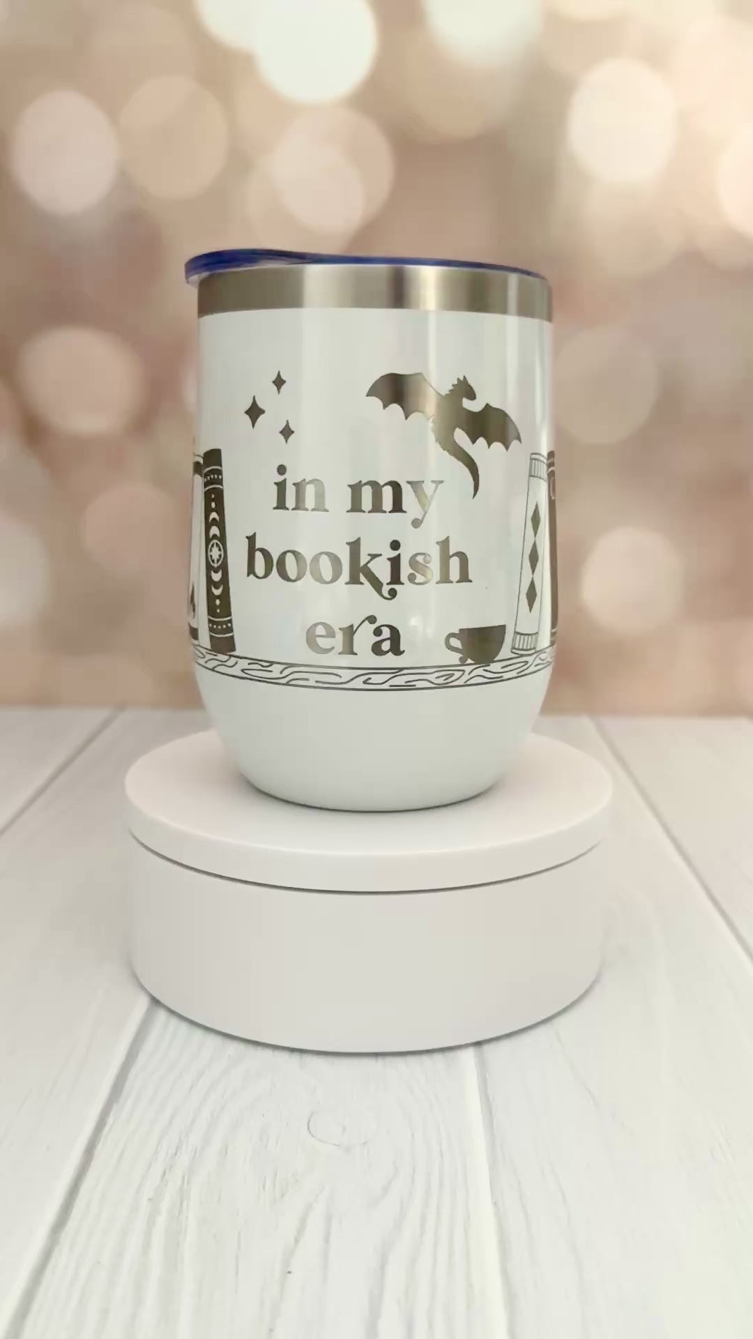 white wine tumbler engraved with "in my bookish era" and books on a bookshelf with a dragon flying above