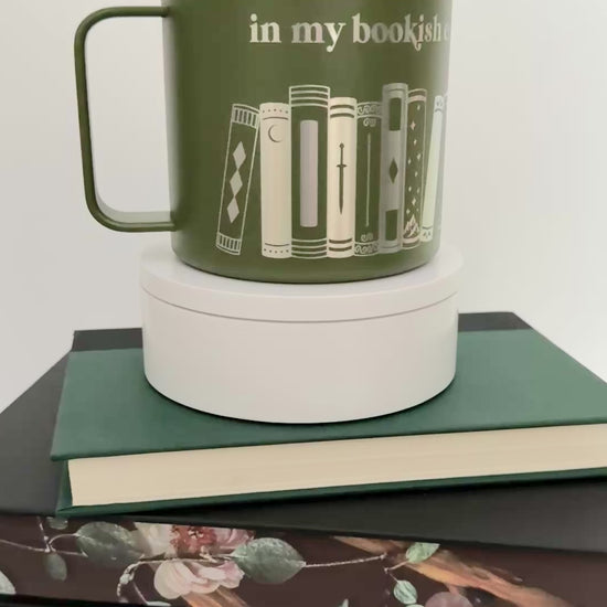 Green stainless steel mug with 'in my bookish era' and books engraved around it, set on top of a stack of green, black and gray books.