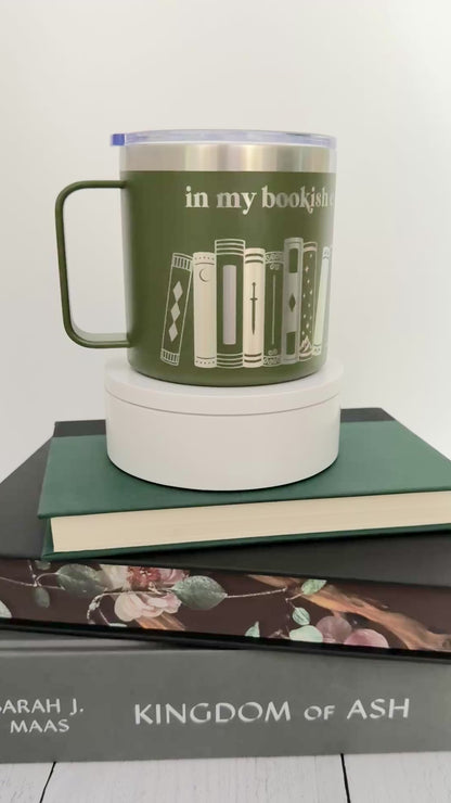 Green stainless steel mug with 'in my bookish era' and books engraved around it, set on top of a stack of green, black and gray books.