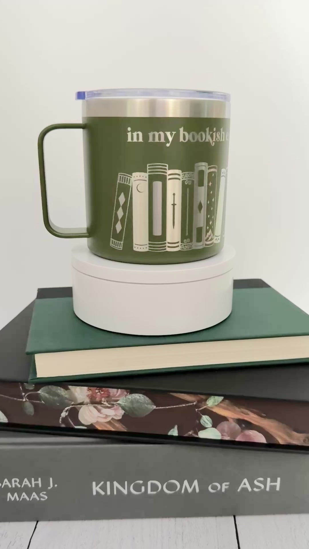 Green stainless steel mug with 'in my bookish era' and books engraved around it, set on top of a stack of green, black and gray books.