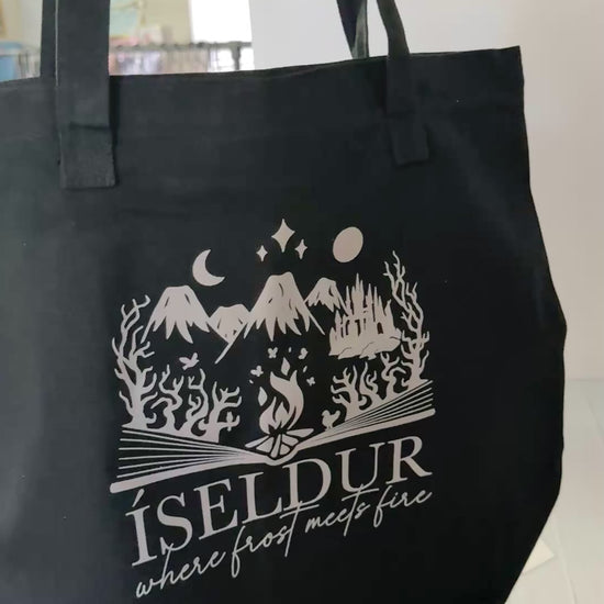 Two sided black tote bag with art inspired by The Road of Bones by Demi Winters, including a bloody axe on a shield and elements from Iseldur spilling from the pages of a book.