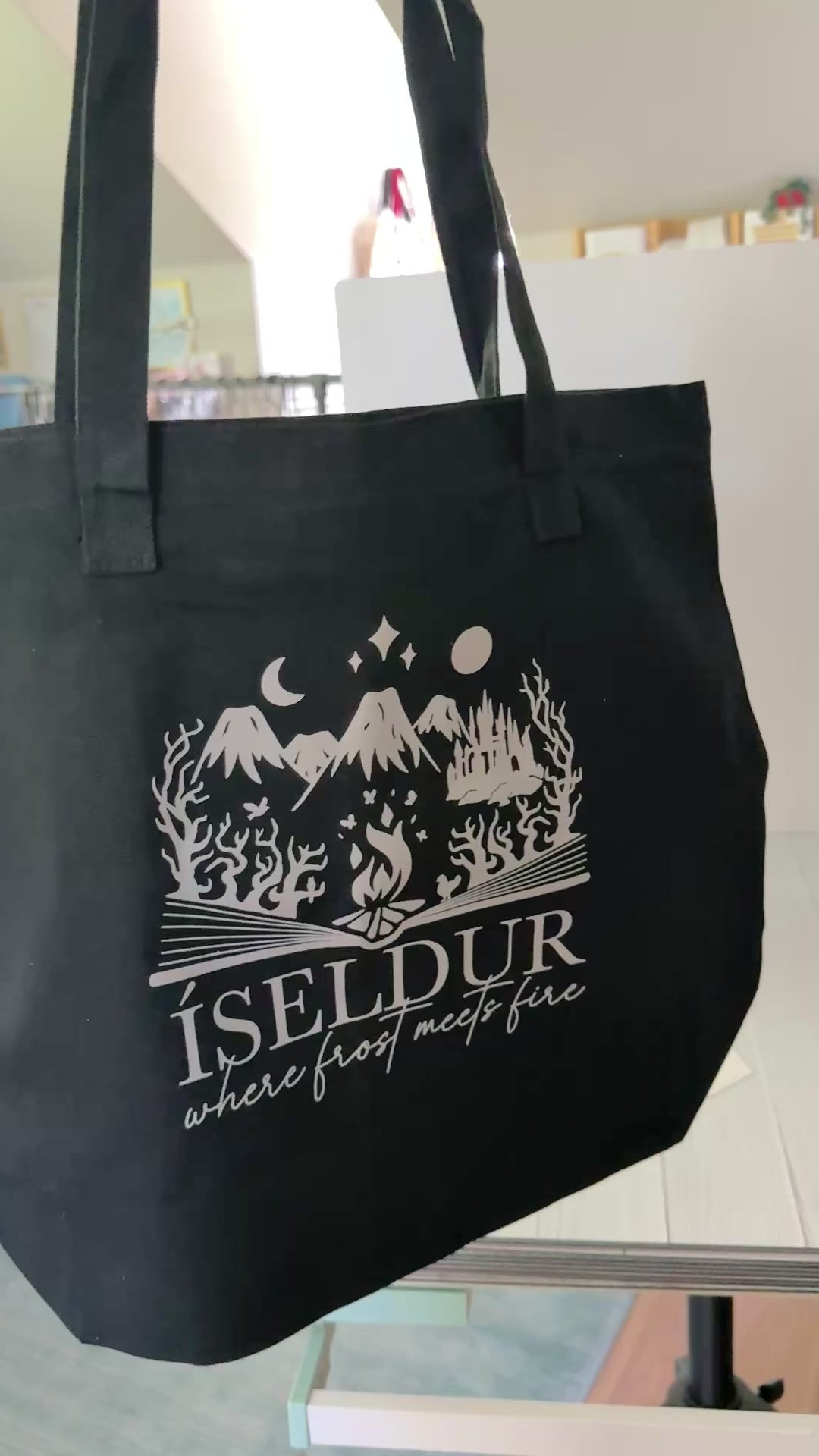 Two sided black tote bag with art inspired by The Road of Bones by Demi Winters, including a bloody axe on a shield and elements from Iseldur spilling from the pages of a book.