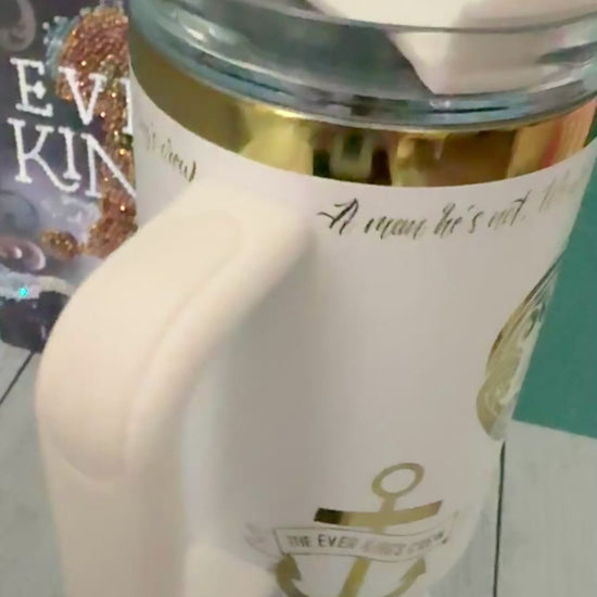 An Ever King themed white handled travel tumbler with a clear lid sits on a white pedestal in front of a teal wall and light wood floor. The tumbler is showing a quote from the book by LJ Andrews: 'a man he's not, we work, we rot, no sleep until it's through. A sailor's grave is all we crave. We are the Ever King's crew.'