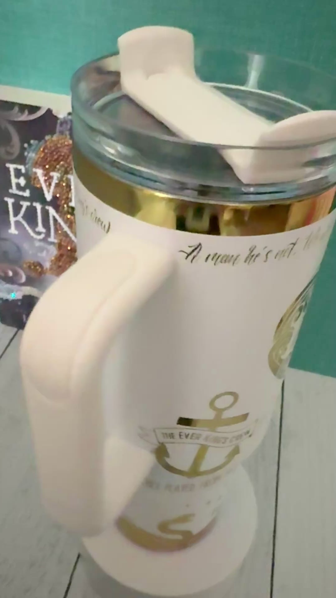 An Ever King themed white handled travel tumbler with a clear lid sits on a white pedestal in front of a teal wall and light wood floor. The tumbler is showing a quote from the book by LJ Andrews: 'a man he's not, we work, we rot, no sleep until it's through. A sailor's grave is all we crave. We are the Ever King's crew.'
