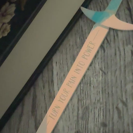 iridescent sword bookmark engraved with 'turn your pain into power' laying in front of 'The Road of Bones' book by Demi Winters