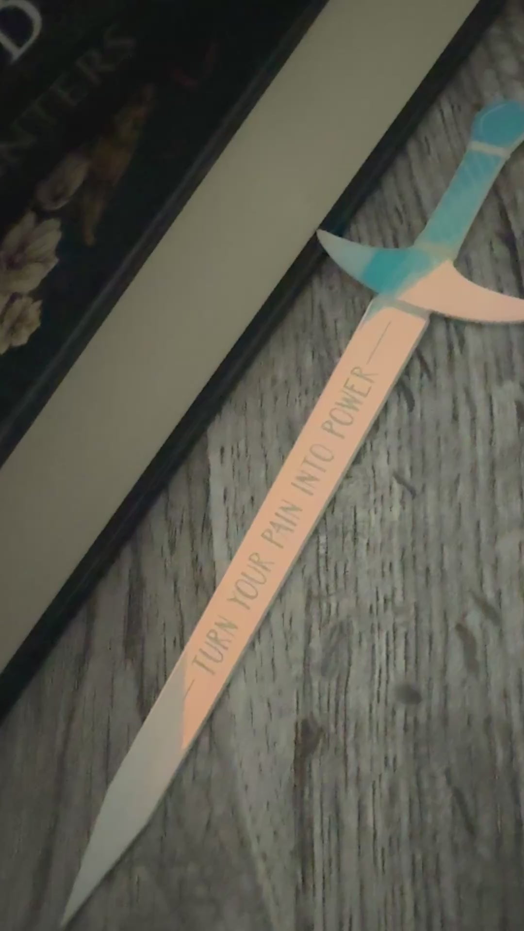iridescent sword bookmark engraved with 'turn your pain into power' laying in front of 'The Road of Bones' book by Demi Winters