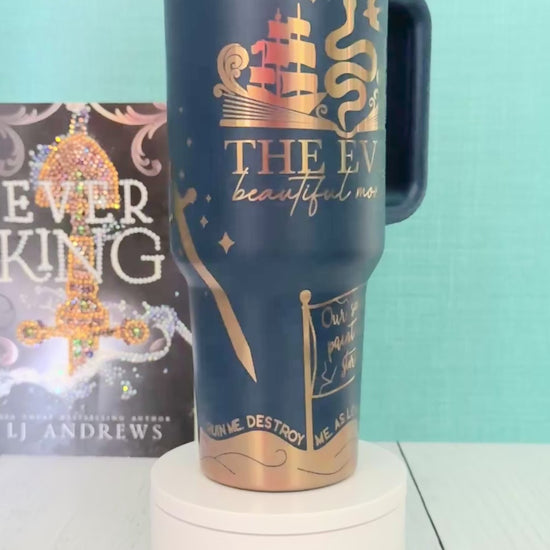 An Ever King themed navy blue handled travel tumbler sits on a white pedestal in front of a teal wall and light wood floor. The tumbler is printed with the title 'The Ever: Beautiful Monster' in script and serif fonts, decorated with a serpent, ship, songbird and gold engraved details.