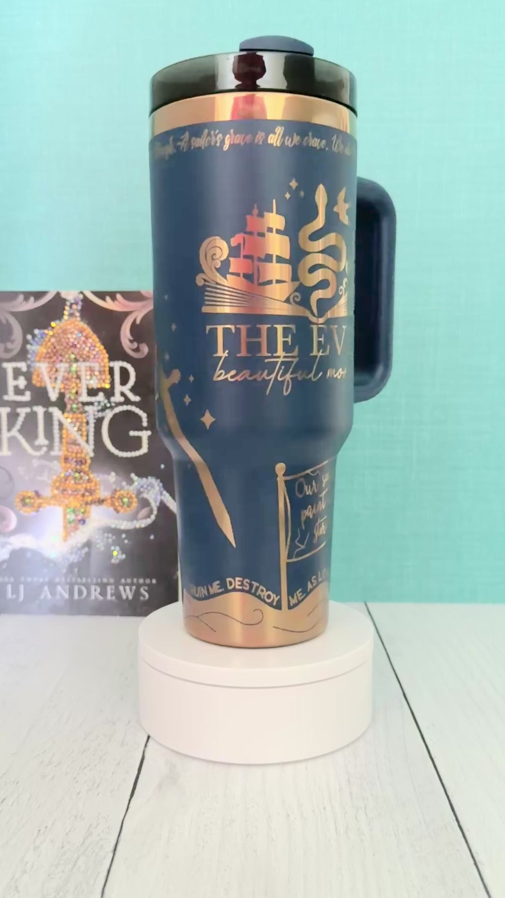 An Ever King themed navy blue handled travel tumbler sits on a white pedestal in front of a teal wall and light wood floor. The tumbler is printed with the title 'The Ever: Beautiful Monster' in script and serif fonts, decorated with a serpent, ship, songbird and gold engraved details.