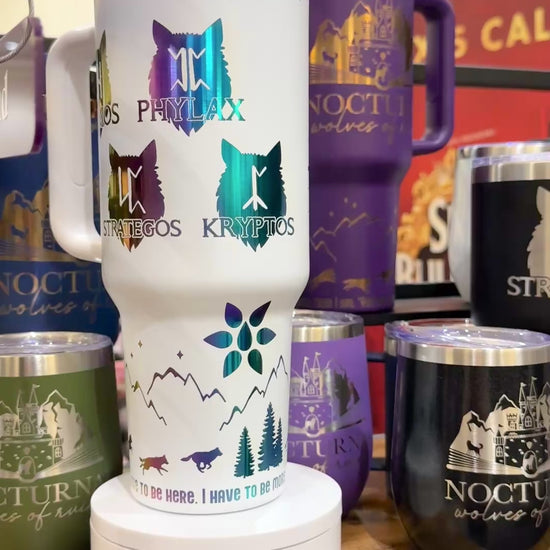 White 40 oz Dire Bound inspired tumbler with rainbow-engraved Nocturna castle, wolf pack symbols for Daemos, Phylax, Strategos and Kryptos, and wolves running around the base of the tumbler with mountains, pine trees and stars and the quote 'you must, so you will.'