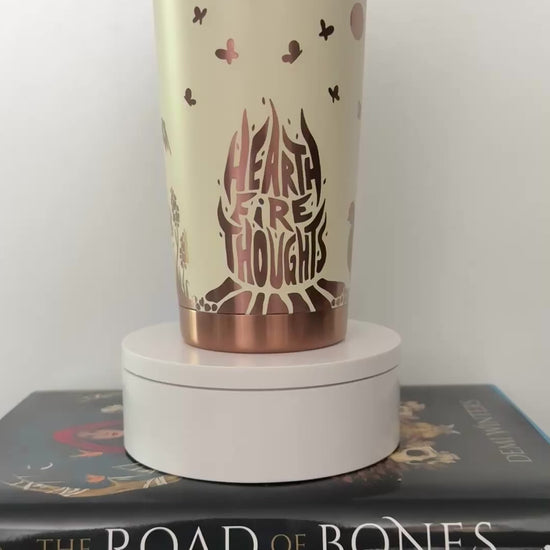 Ivory tumbler engraved with gold designs inspired by Demi Winters' Ashen series, spinning on top of a set of the books, including Road of Bones and Kingdom of Claw.