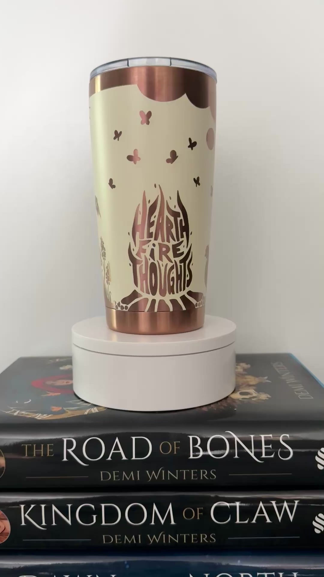Ivory tumbler engraved with gold designs inspired by Demi Winters' Ashen series, spinning on top of a set of the books, including Road of Bones and Kingdom of Claw.