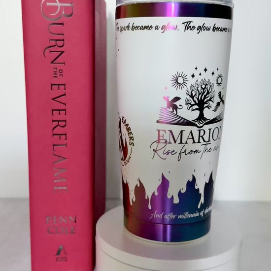 Slow turn of a white and rainbow iridescent 20 oz Emarion tumbler with Everflame tree, phoenix and gryvern exploding from an open book and other world elements from  the Kindred's Curse Saga, next to a copy of Burn of the Everflame