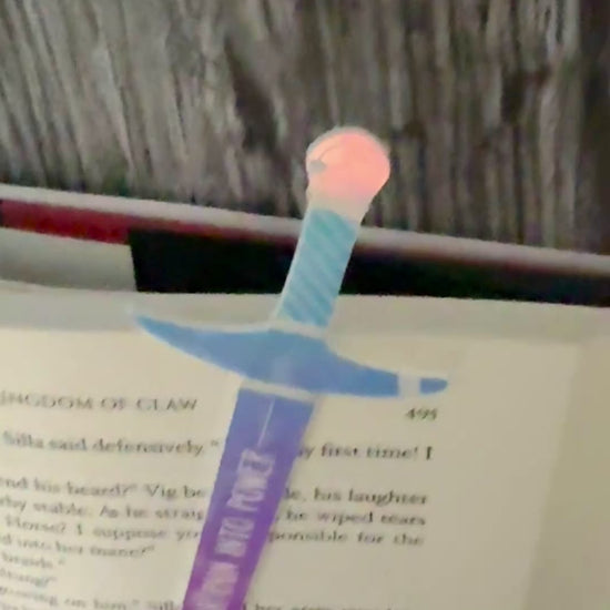 iridescent sword bookmark engraved with 'turn your pain into power' laying on an open book 'The Road of Bones' by Demi Winters