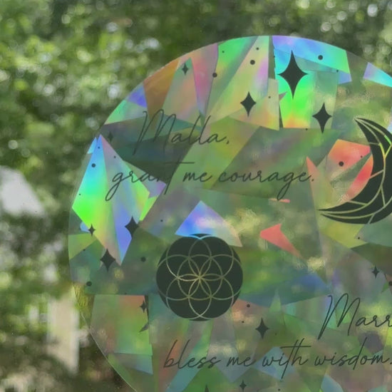 Decorative circular suncatcher inspired by The Road of Bones by Demi Winters with text 'Malla grant me courage. Marra bless me with wisdom' and moon and stars symbols on a window with a blurred outdoor background.
