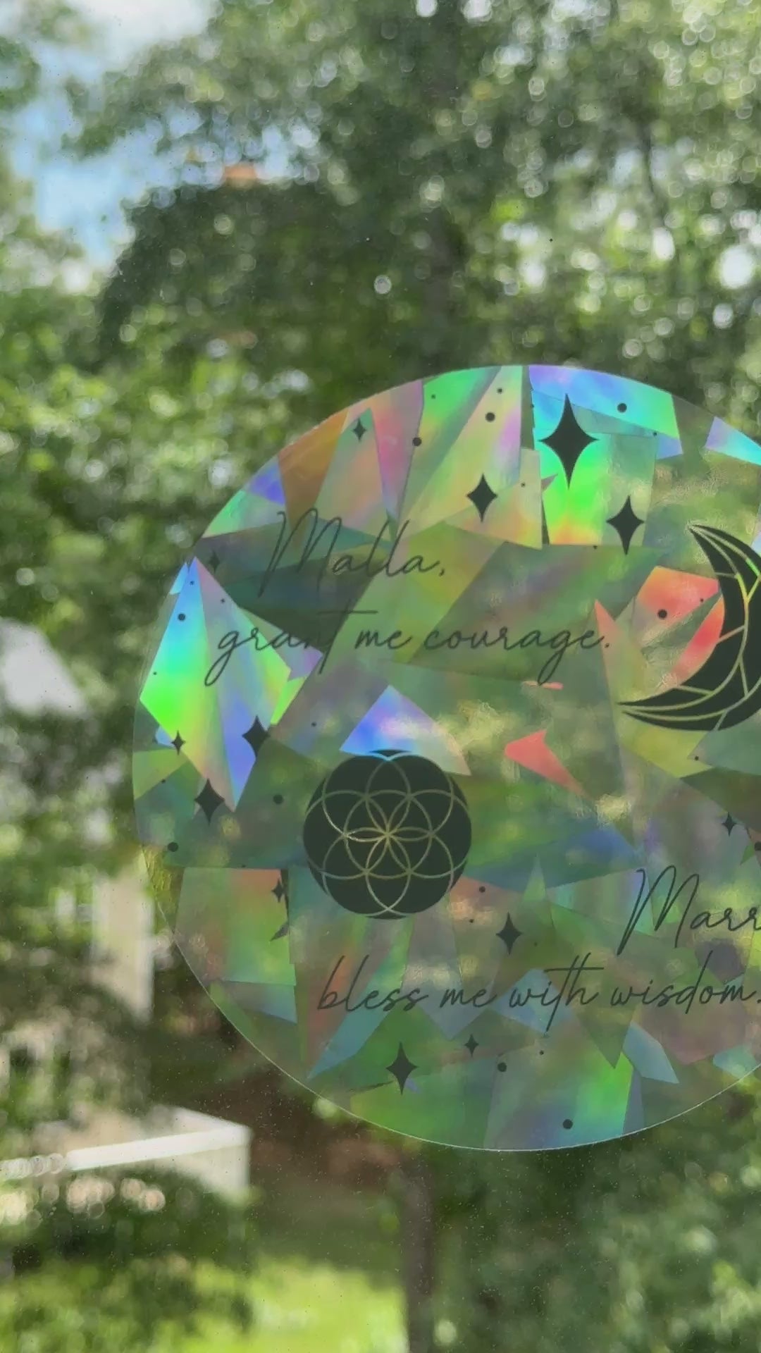 Decorative circular suncatcher inspired by The Road of Bones by Demi Winters with text 'Malla grant me courage. Marra bless me with wisdom' and moon and stars symbols on a window with a blurred outdoor background.