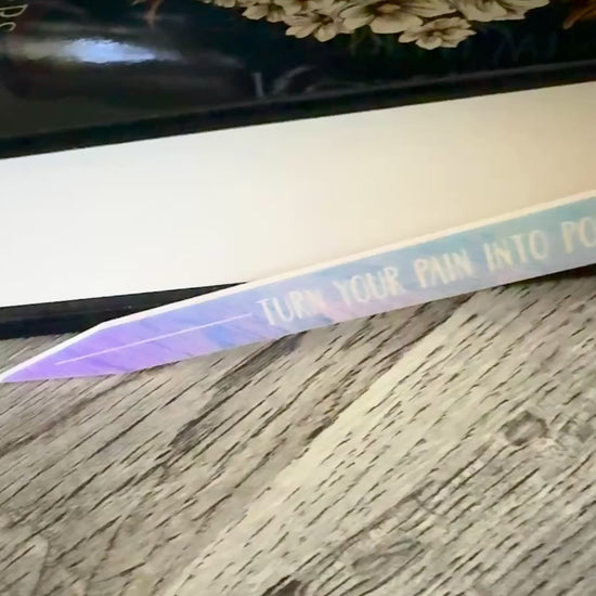 iridescent sword bookmark engraved with 'turn your pain into power' laying in front of 'The Road of Bones' book by Demi Winters