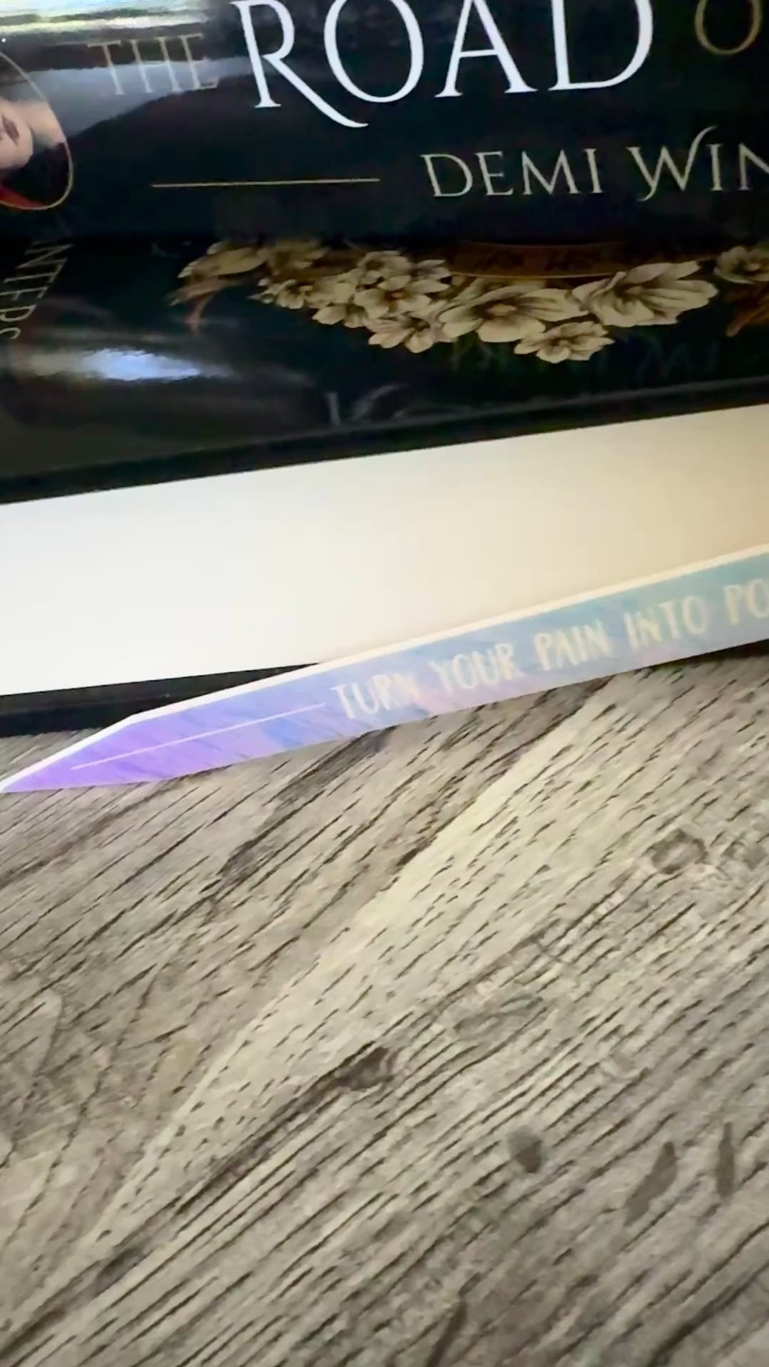 iridescent sword bookmark engraved with 'turn your pain into power' laying in front of 'The Road of Bones' book by Demi Winters