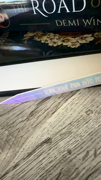 iridescent sword bookmark engraved with 'turn your pain into power' laying in front of 'The Road of Bones' book by Demi Winters