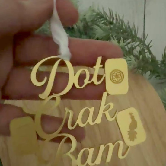 A gold metallic mahjong Christmas ornament spelling out 'Dot Crak Bam' with traditional mahjong tiles, hanging from a white ribbon on a wooden surface with green pine branches in the background.