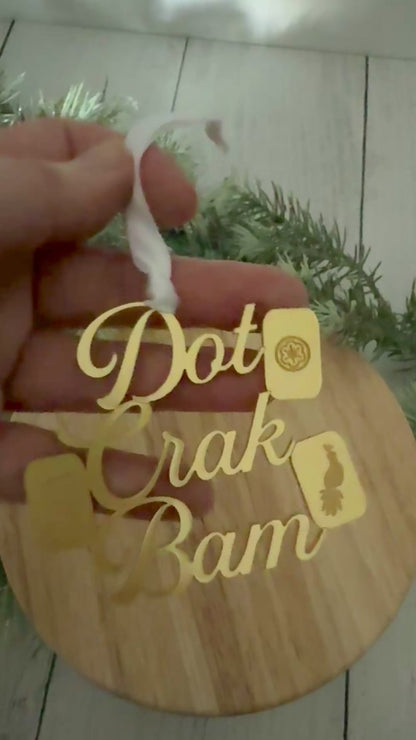 A gold metallic mahjong Christmas ornament spelling out 'Dot Crak Bam' with traditional mahjong tiles, hanging from a white ribbon on a wooden surface with green pine branches in the background.
