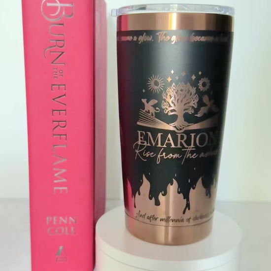 Slow turn of a black and copper 20 oz Emarion tumbler with Everflame tree, phoenix and gryvern exploding from an open book and other world elements from  the Kindred's Curse Saga, next to a copy of Burn of the Everflame