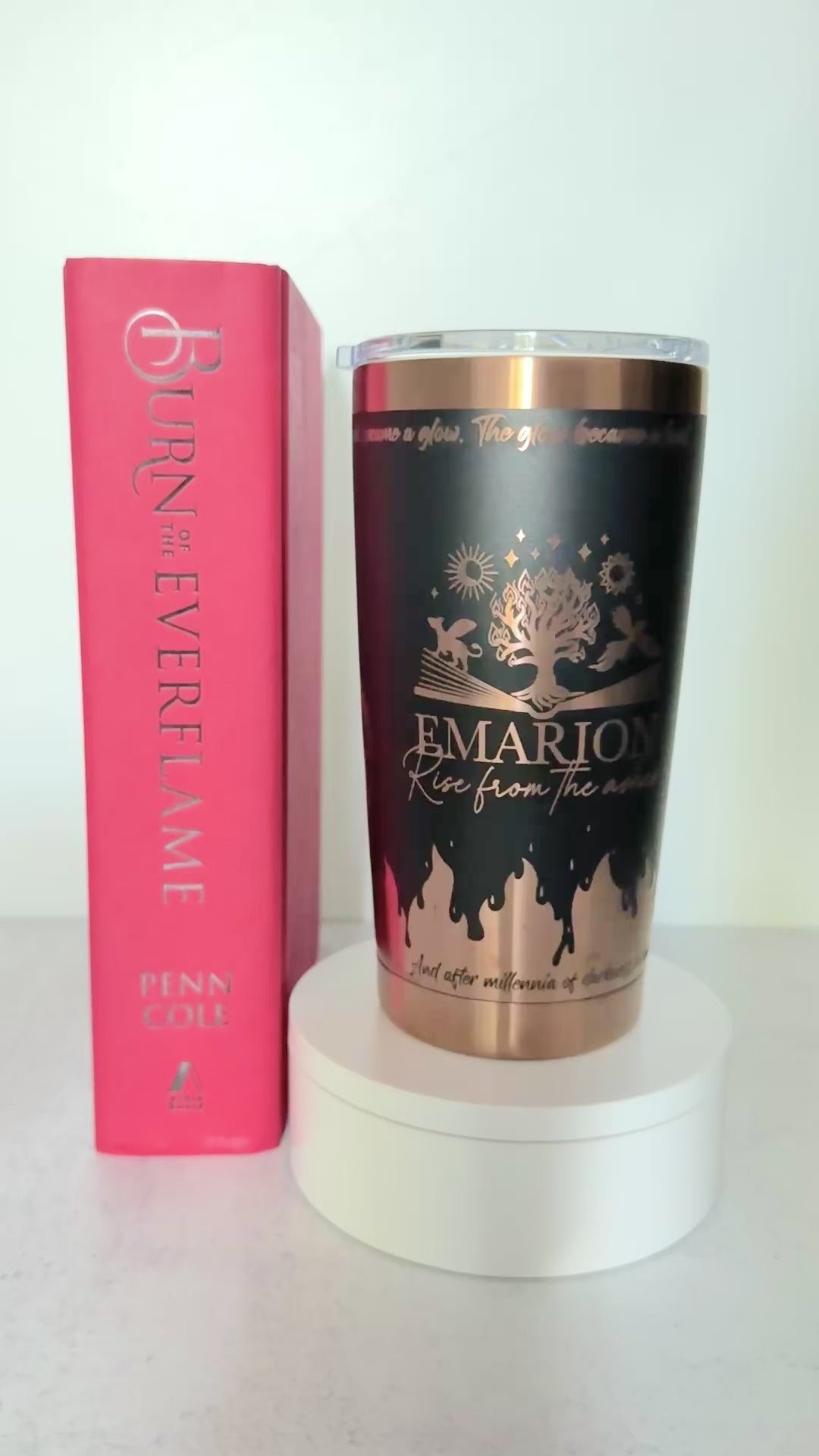 Slow turn of a black and copper 20 oz Emarion tumbler with Everflame tree, phoenix and gryvern exploding from an open book and other world elements from  the Kindred's Curse Saga, next to a copy of Burn of the Everflame