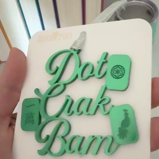 A green metallic mahjong Christmas ornament spelling out 'Dot Crak Bam' with traditional mahjong tiles, hanging from a silver ribbon on a white card.