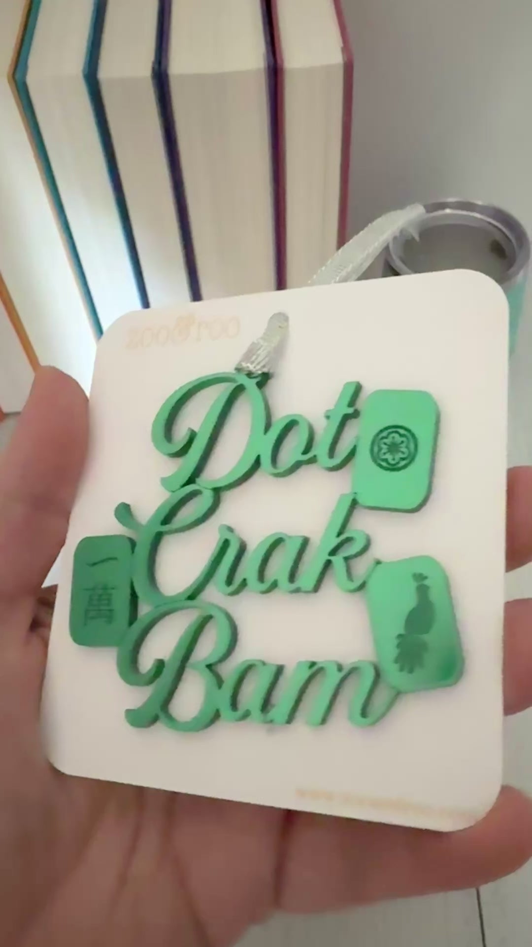 A green metallic mahjong Christmas ornament spelling out 'Dot Crak Bam' with traditional mahjong tiles, hanging from a silver ribbon on a white card.