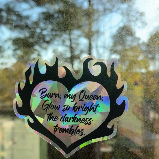 Flaming heart suncatcher decal on a window with the quote "Burn, my Queen. Glow so bright the darkness trembles."