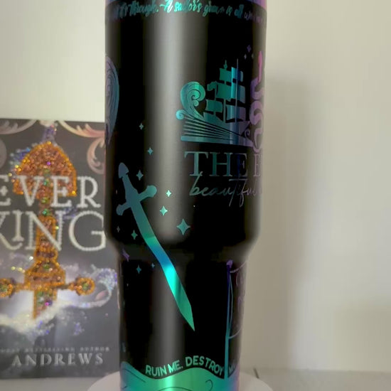 A black Ever King themed tumbler with rainbow engraved details from the books by LJ Andrews including a heart made of waves that says 'hello love' and an open book design with ship, serpent and songbird that says 'the Ever: beautiful monster'