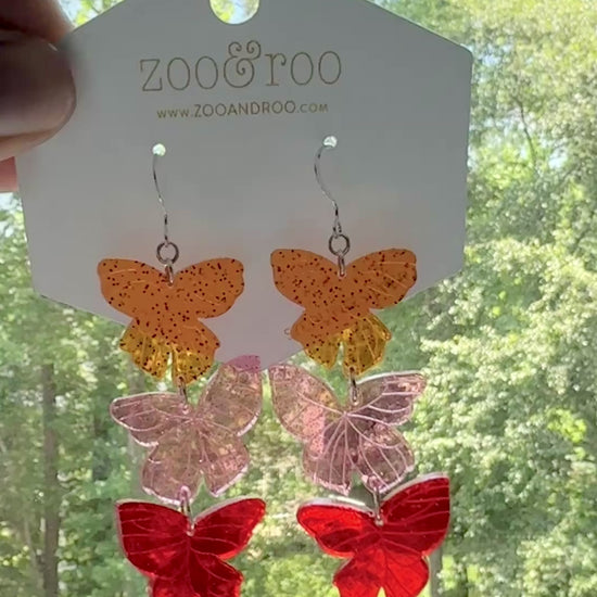 Flíta butterfly earrings inspired by Road of Bones by Demi Winters on a display card with zoo&roo branding, against a window with tree background.