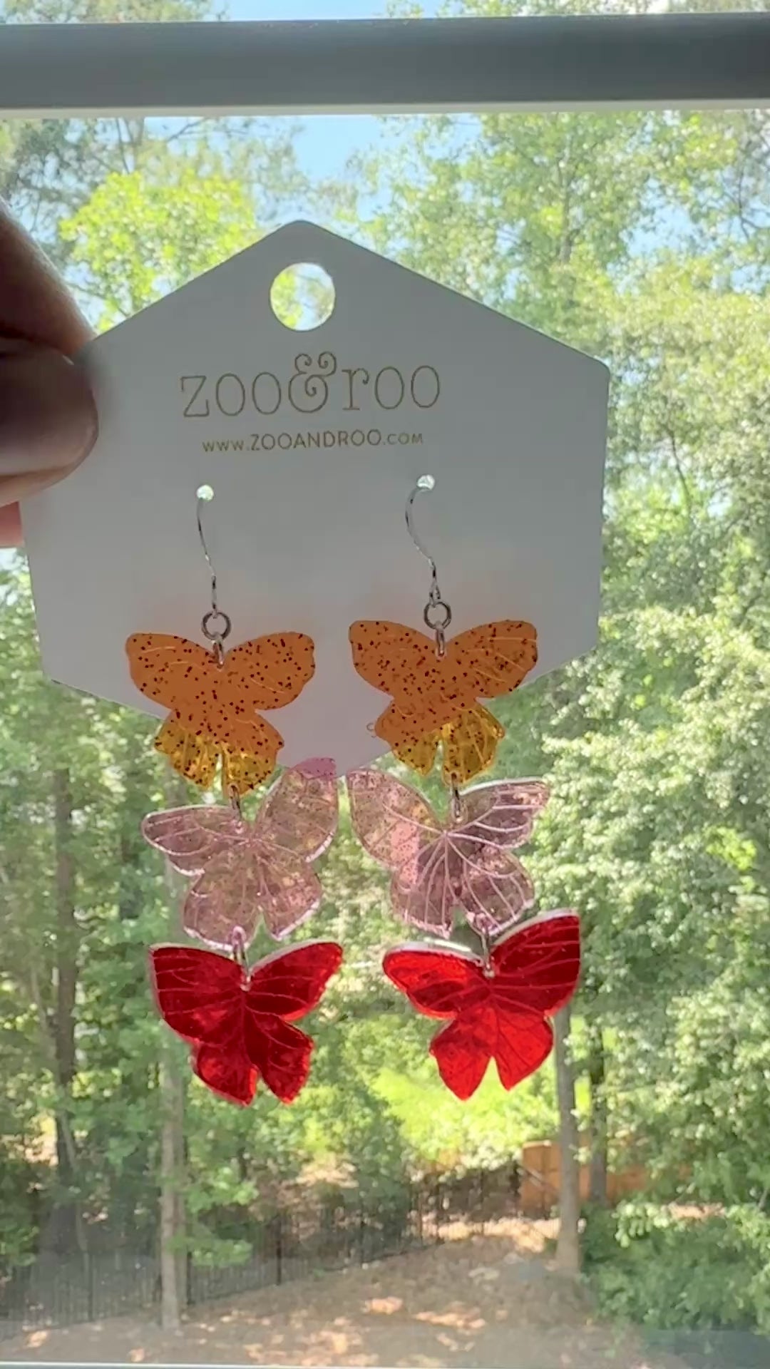 Flíta butterfly earrings inspired by Road of Bones by Demi Winters on a display card with zoo&roo branding, against a window with tree background.