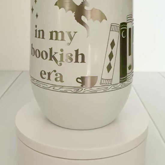 white wine tumbler spinning around, engraved with "in my bookish era" and books on a book shelf with a dragon