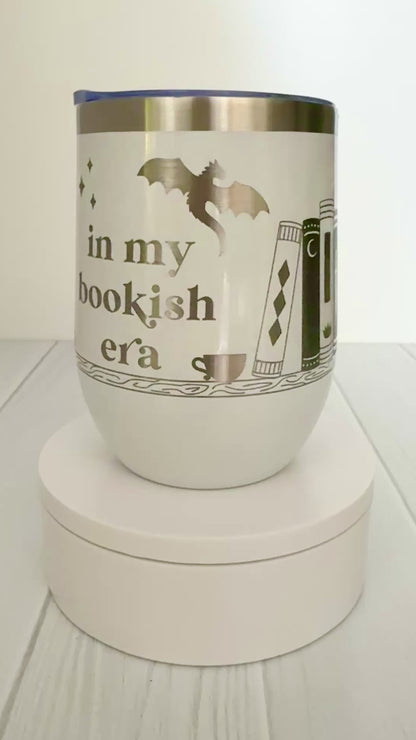 white wine tumbler spinning around, engraved with "in my bookish era" and books on a book shelf with a dragon