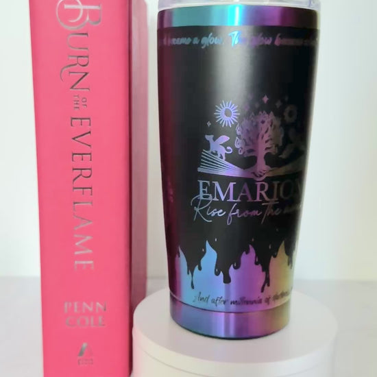 Slow turn of a black and rainbow iridescent 20 oz Emarion tumbler with Everflame tree, phoenix and gryvern exploding from an open book and other world elements from  the Kindred's Curse Saga, next to a copy of Burn of the Everflame