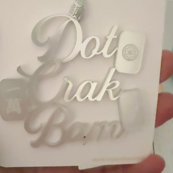 A silver metallic mahjong Christmas ornament spelling out 'Dot Crak Bam' with traditional mahjong tiles, hanging from a silver ribbon on a white card.