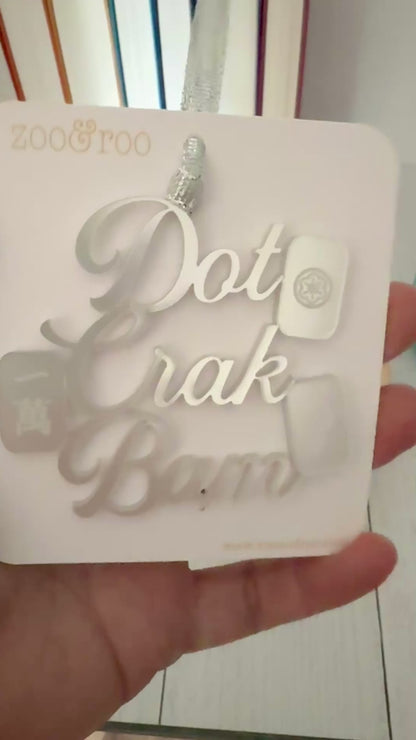 A silver metallic mahjong Christmas ornament spelling out 'Dot Crak Bam' with traditional mahjong tiles, hanging from a silver ribbon on a white card.